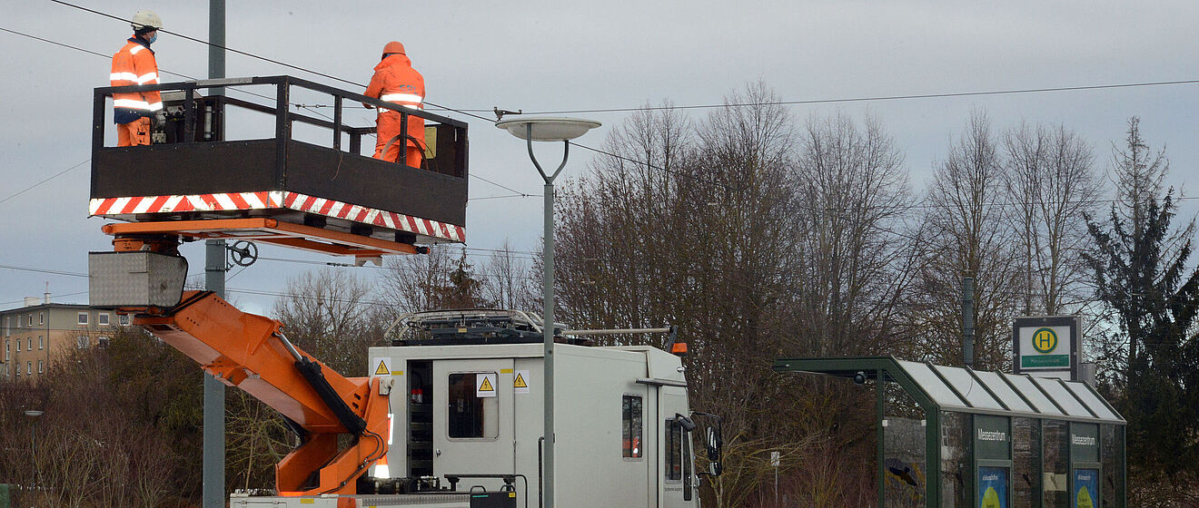 Zwei Männer in orangener Schutzkleidung stehen auf einem Personenaufzug und arbeiten an einer Stromoberleitung. Im Hintergrund ist eine Haltestelle.