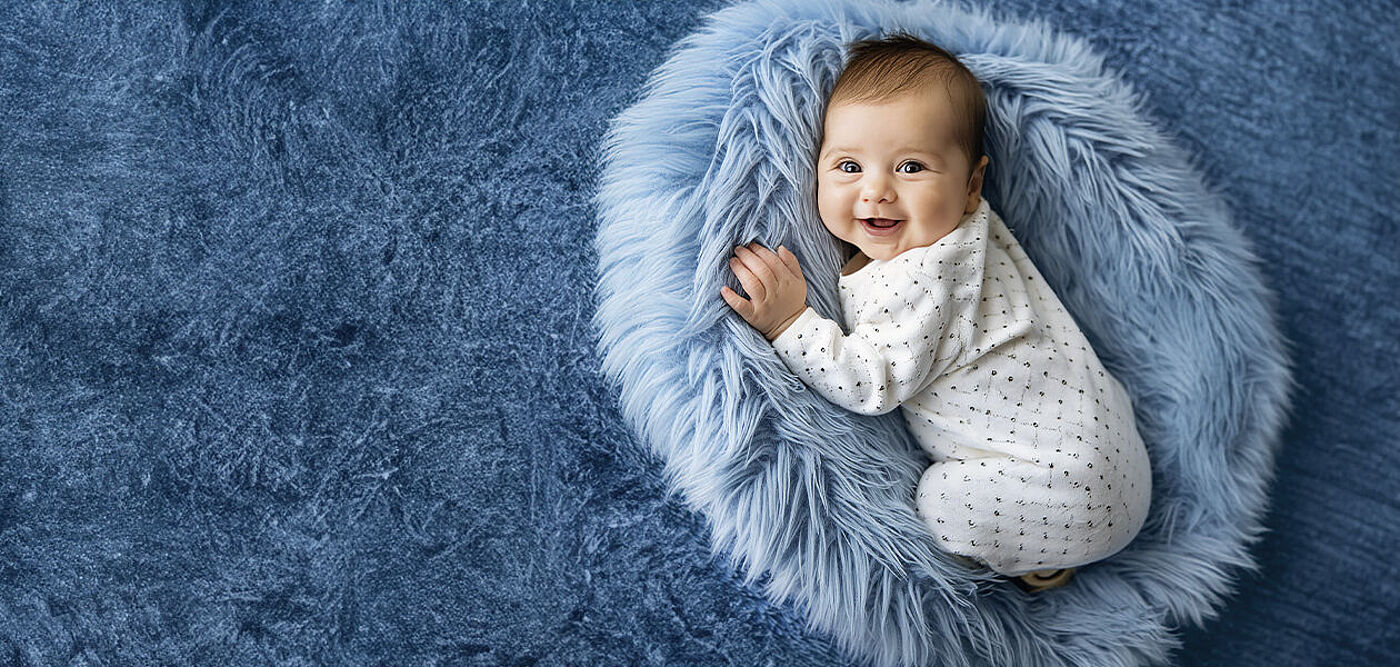 Ein lachendes Baby liegt auf einem grauen Wollkissen und schaut in die Kamera