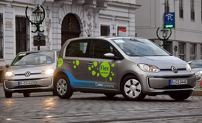 Carsharing Autos fahren auf einer gepflasterten Straße, Weiterleitung zu Überblick Preise und Tarife