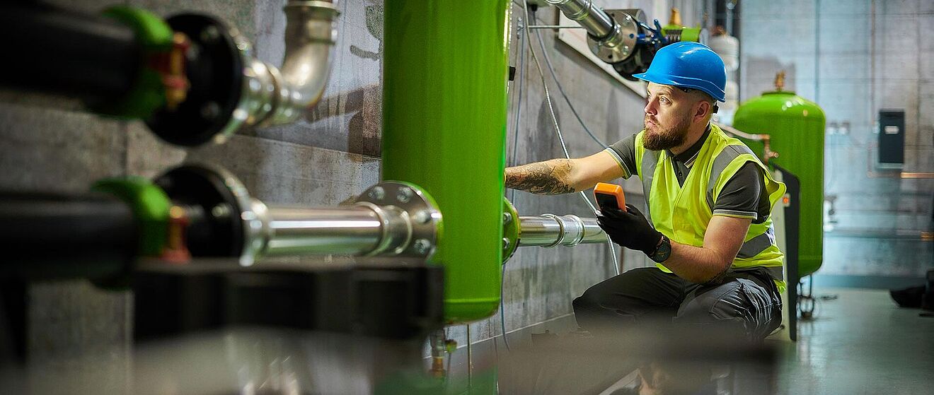 Mann mit Helm, der Anlagen überprüft und ein Messgerät hält. Weiterverlinkung zu Energiedienstleistungsseite