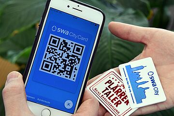 Eine Hand hält ein Smartphone mit einem QR-Code der swa CityCard auf dem Bildschirm. Die recht Hand hält eine physische swa City Karte und einen Plärrertaler